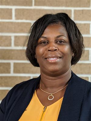 Principal Wilson wearing a yellow shirt and black jacket standing in front of brown brick wall. 